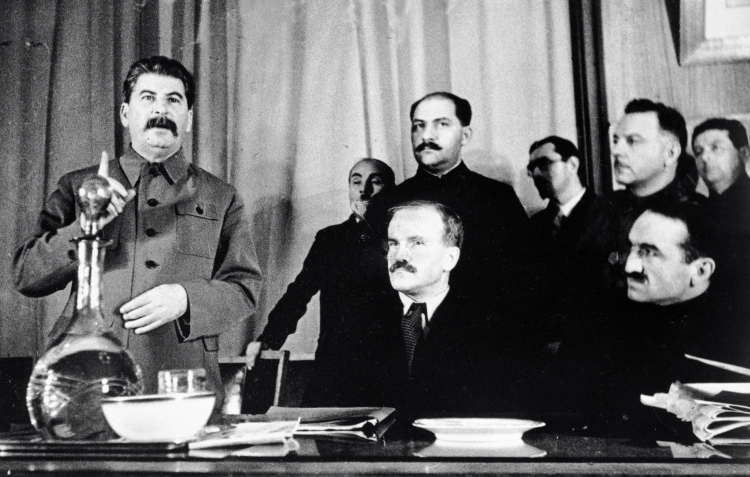 Стоят в первом ряду слева направо: Иосиф Сталин, Лазарь Каганович, Климент Ворошилов. Сидят слева направо: Вячеслав Молотов, Анастас Микоян.