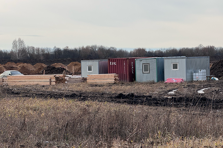 На стройплощадке видно около 10 человек, многочисленные грузовики и тракторы, также готовы вагончики для строителей