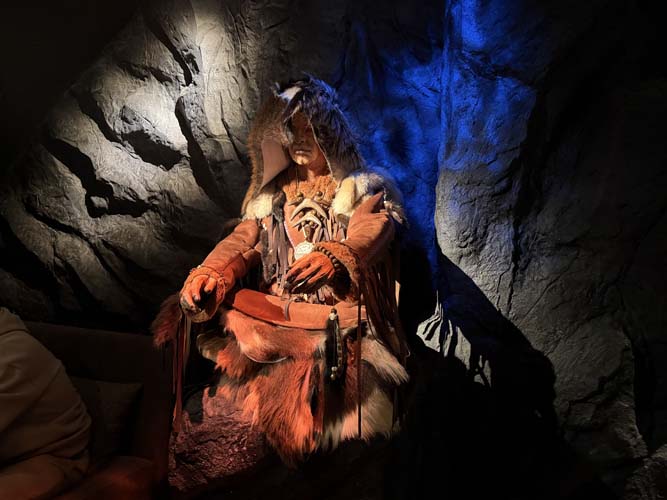 Шаман в полном ритуальном облачении: капюшон с перьями, слои кожи и меха, бахрома, амулет на груди, рыжие меховые рукава, на поясе &mdash; юбка из лисьих хвостов